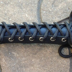 May include: Close-up of a black leather handle with a woven design. The handle is wrapped with black cord, laced through silver eyelets. The handle is part of a larger object, with a portion of the object visible on the right side.