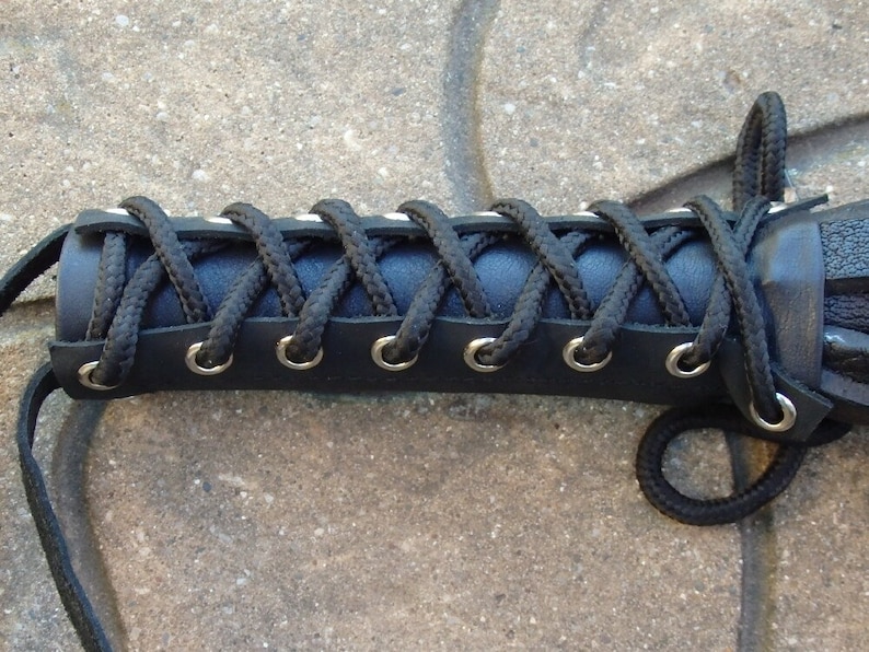 May include: Close-up of a black leather handle with a woven design. The handle is wrapped with black cord, laced through silver eyelets. The handle is part of a larger object, with a portion of the object visible on the right side.