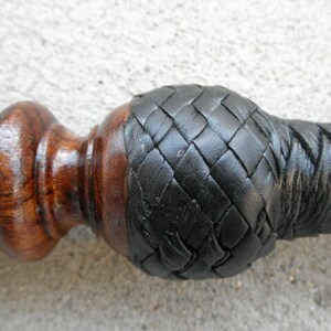 May include: A close-up of a wooden walking stick with a black leather braided handle. The handle is wrapped in a diamond pattern.