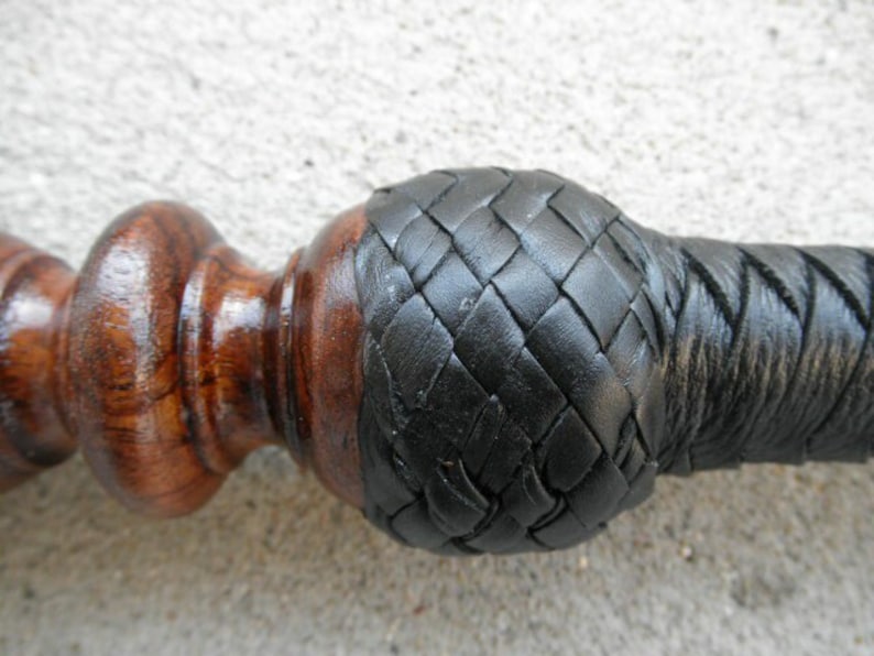 May include: A close-up of a wooden walking stick with a black leather braided handle. The handle is wrapped in a diamond pattern.