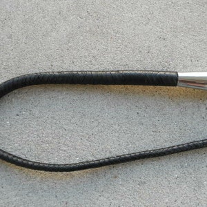 May include: A black leather riding crop with a braided handle and a polished silver metal handle. The crop is curved and lies on a gray surface. The handle is smooth and rounded.