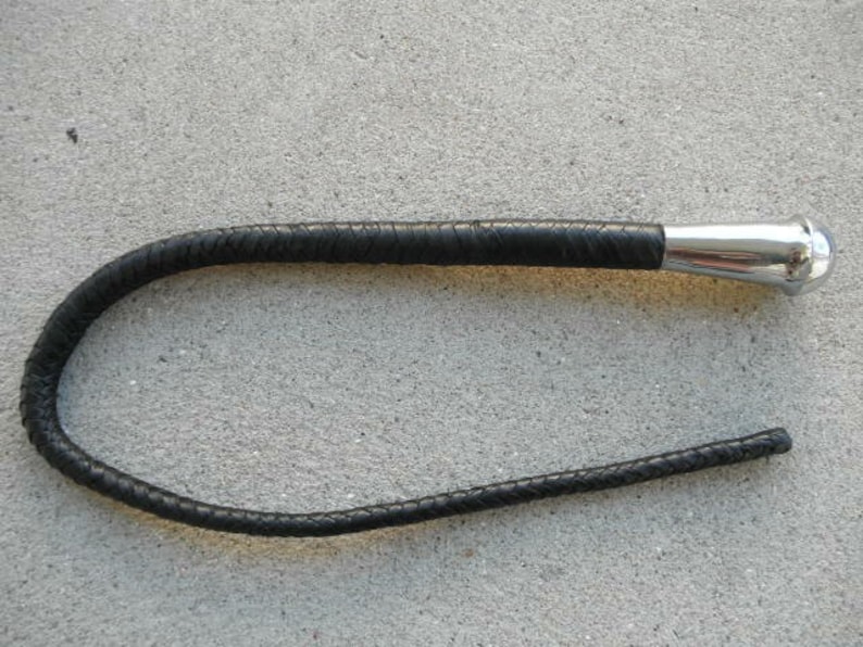 May include: A black leather riding crop with a braided handle and a polished silver metal handle. The crop is curved and lies on a gray surface. The handle is smooth and rounded.