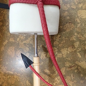 May include: A red leather whip with a braided handle and a black arrowhead-shaped tip. The whip is attached to a white mannequin torso. The handle is made of wood and metal.