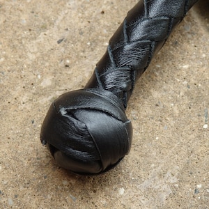 May include: Close-up of a black leather whip. The handle is tightly braided, transitioning into a rounded, stitched end. The leather appears smooth and well-maintained, with a slight sheen. The whip is set against a textured, light-colored surface.