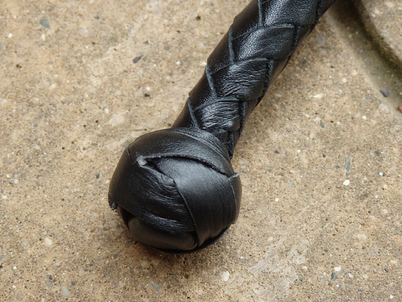 May include: Close-up of a black leather whip. The handle is tightly braided, transitioning into a rounded, stitched end. The leather appears smooth and well-maintained, with a slight sheen. The whip is set against a textured, light-colored surface.