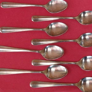 Puede incluir: Ocho cucharas de plata dispuestas en fila sobre un fondo rojo. Las cucharas son todas del mismo tamaño y forma, con un cuenco redondeado y un mango largo y delgado.