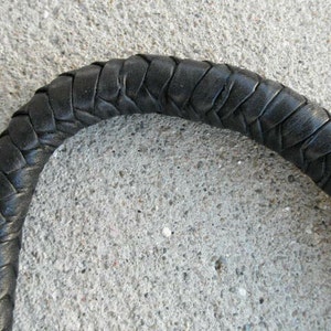 May include: Close-up of a black, braided leather handle. The handle is curved and appears to be part of a larger object. The leather is tightly woven, creating a textured surface. The background is a rough, gray concrete.