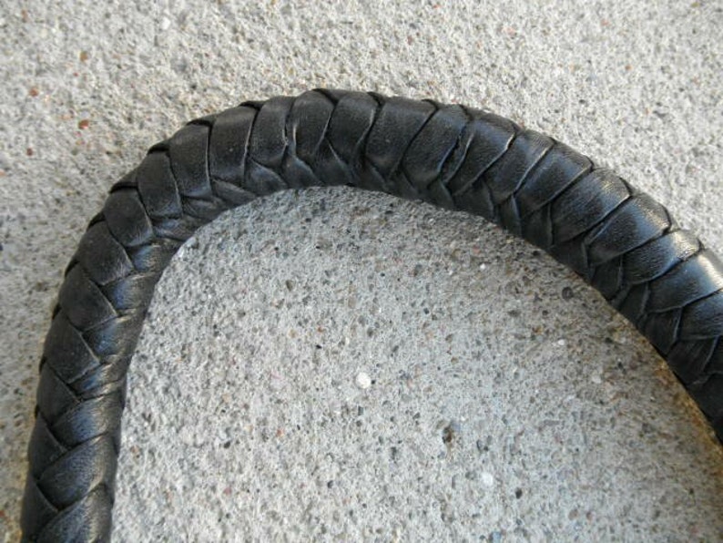 May include: Close-up of a black, braided leather handle. The handle is curved and appears to be part of a larger object. The leather is tightly woven, creating a textured surface. The background is a rough, gray concrete.