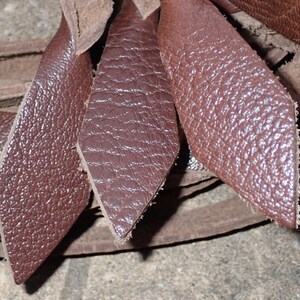 May include: Close-up of several diamond-shaped pieces of brown leather. The leather has a textured surface and appears to be cut from a larger piece. The edges of the leather are rough, and the pieces are overlapping.