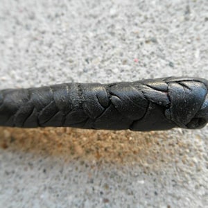 May include: Close-up of a black leather whip. The whip is tightly braided, with a rounded end. The leather appears worn, with visible texture and slight imperfections. The background is a rough, gray surface.