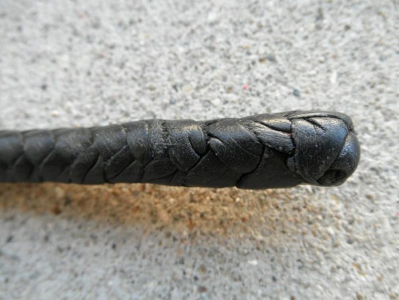 May include: Close-up of a black leather whip. The whip is tightly braided, with a rounded end. The leather appears worn, with visible texture and slight imperfections. The background is a rough, gray surface.