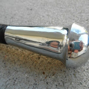 May include: Close-up of a riding crop with a black leather handle and a polished chrome head. The chrome reflects the surroundings, showing a distorted image. The handle appears to be braided.