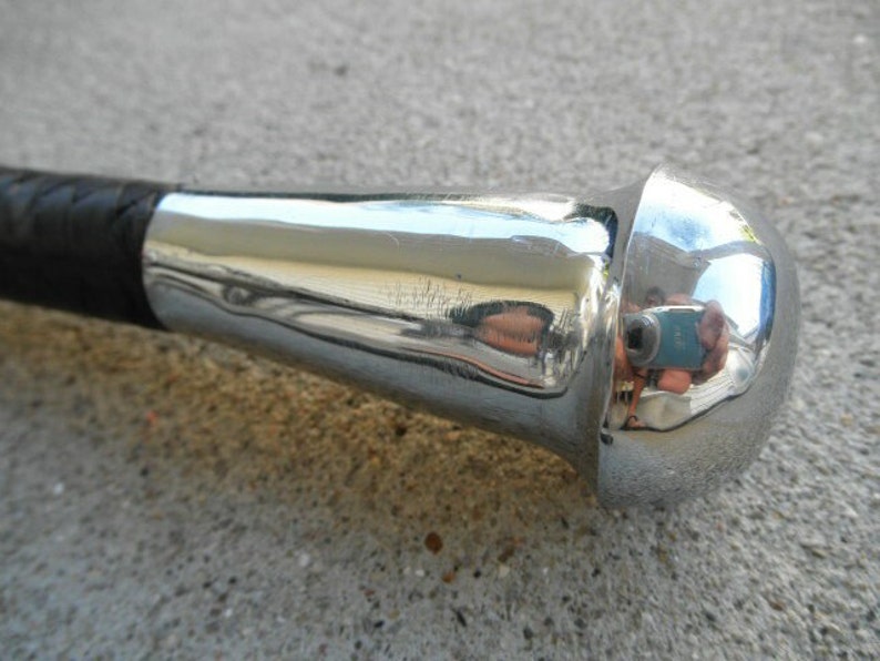 May include: Close-up of a riding crop with a black leather handle and a polished chrome head. The chrome reflects the surroundings, showing a distorted image. The handle appears to be braided.