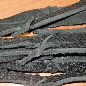 May include: Close-up of several black leather strips with a textured surface. The strips are layered and tied together with smaller leather pieces. The leather appears to be soft and pliable, with a matte finish.