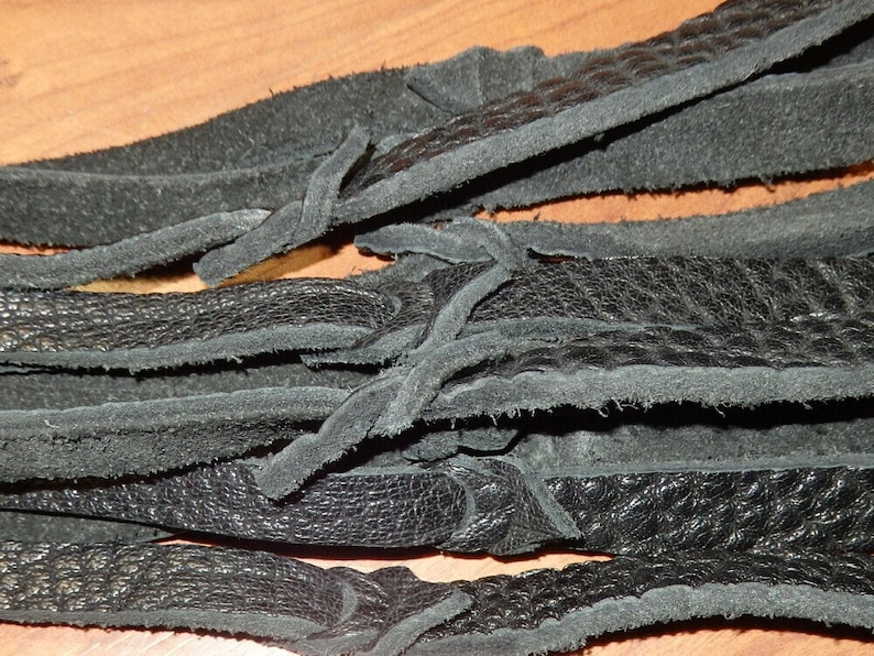 May include: Close-up of several black leather strips with a textured surface. The strips are layered and tied together with smaller leather pieces. The leather appears to be soft and pliable, with a matte finish.