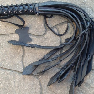 May include: A black leather whip with a braided handle and multiple long, flat strands. The handle is laced with black cord and has a small leather tag attached. The whip is designed for a specific purpose.