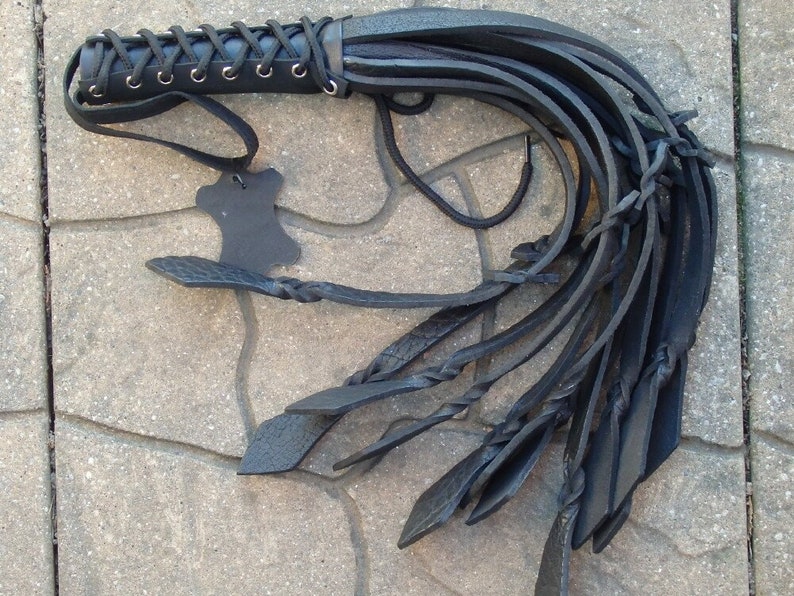 May include: A black leather whip with a braided handle and multiple long, flat strands. The handle is laced with black cord and has a small leather tag attached. The whip is designed for a specific purpose.