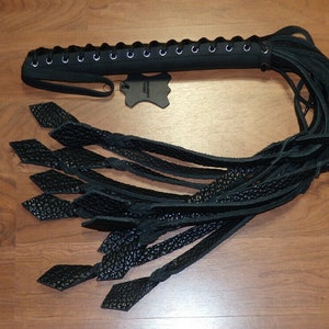 May include: A black leather whip with a braided handle and multiple long, textured leather strands. The handle is laced with a series of eyelets. The ends of the strands have diamond-shaped accents. The whip is displayed on a wooden surface.
