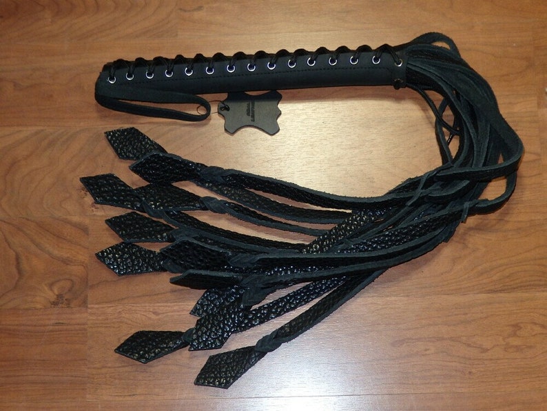 May include: A black leather whip with a braided handle and multiple long, textured leather strands. The handle is laced with a series of eyelets. The ends of the strands have diamond-shaped accents. The whip is displayed on a wooden surface.