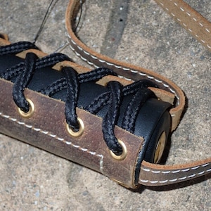 May include: Close-up of a handcrafted leather item with a dark brown and black color scheme. The item features a black cord laced through brass eyelets, with a light brown leather strap and white stitching.