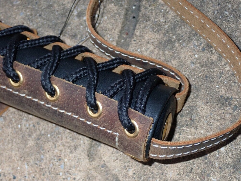 May include: Close-up of a handcrafted leather item with a dark brown and black color scheme. The item features a black cord laced through brass eyelets, with a light brown leather strap and white stitching.