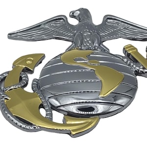 May include: A silver and gold emblem featuring an eagle perched atop a globe with gold continents, an anchor, and the letters USMC. The emblem is a symbol of the United States Marine Corps.
