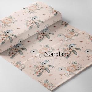 May include: A roll of fabric with a repeating pattern of sea turtles decorated with flowers. The turtles are in grey, beige, and blue, set against a light pink background with small floral accents. The fabric has the text "NoirBlanc" printed on it.