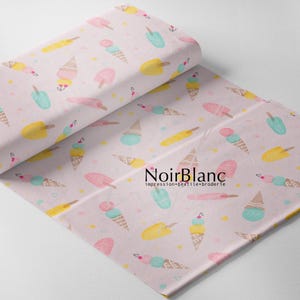 May include: A roll of light pink fabric featuring a repeating pattern of colorful ice cream cones and popsicles in shades of yellow, teal, and pink, with the text NoirBlanc impression textile broderie printed in black on the fabric surface.