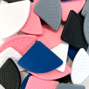 May include: Assortment of silicone baby teethers in various colors and shapes, including pink, blue, white, gray, and black. The teethers feature a variety of textures and patterns, including hearts, dots, and hexagons.