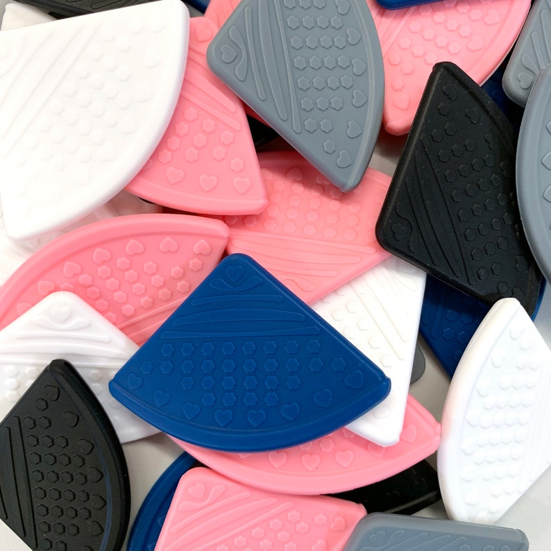 May include: Assortment of silicone baby teethers in various colors and shapes, including pink, blue, white, gray, and black. The teethers feature a variety of textures and patterns, including hearts, dots, and hexagons.