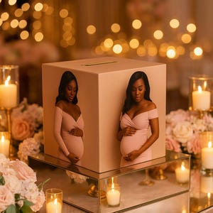 May include: A blush-coloured box with a slot on top, featuring images of a pregnant person in a pink dress. The box sits on a mirrored surface, surrounded by candles and floral arrangements, creating a warm, celebratory atmosphere.