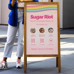 May include: A wooden A-frame sign with a pink background featuring the text Sugar Riot, Detroit's most photogenic dessert truck, and Featured In. It displays images of cookie dough, mini donuts, and beignets, along with three QR codes and social media links.
