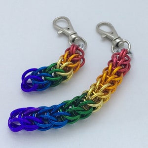 May include: Two rainbow-coloured chainmail keychains. The keychains are made of small, interlocking metal rings in a gradient of colours, including red, orange, yellow, green, blue, and purple. Each keychain has a silver-coloured clasp.