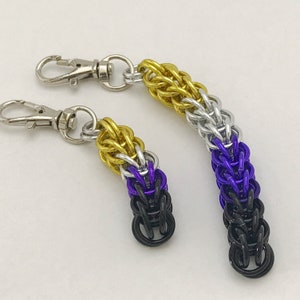 May include: Two keychain charms with a chainmail design in the colours of the non-binary pride flag: yellow, white, purple, and black.
