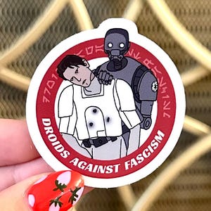 Puede incluir: Una pegatina redonda con borde rojo que presenta una ilustración temática de Star Wars. La imagen representa un droide y un personaje humano, con el texto "DROIDS AGAINST FASCISM" y otro texto en círculo.