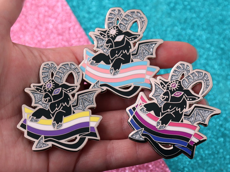 May include: Three black and gold enamel pins featuring a cartoon goat with wings and horns. Each goat is holding a different pride flag: transgender, non-binary, and bisexual.