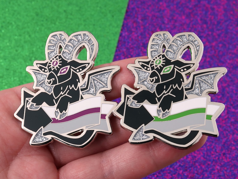 May include: Two black and silver enamel pins featuring a black goat with wings and horns holding a banner with the asexual pride flag colors. The goat has a third eye with a purple iris and a green iris in the other eye. The banner has a white background with a purple stripe, a gray stripe, and a green stripe.