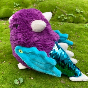 Pode incluir: Brinquedo de pel&uacute;cia de drag&atilde;o roxo e azul com uma cauda verde brilhante e um chifre branco. O drag&atilde;o tem um olho verde e um corpo felpudo.