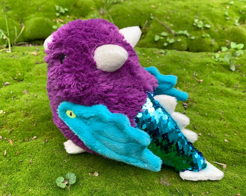 Pode incluir: Brinquedo de pel&uacute;cia de drag&atilde;o roxo e azul com uma cauda verde brilhante e um chifre branco. O drag&atilde;o tem um olho verde e um corpo felpudo.