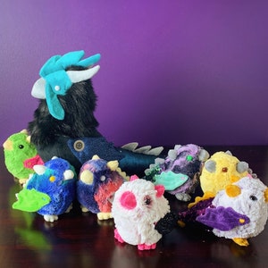 Pode incluir: Um grupo de brinquedos de pel&uacute;cia, incluindo um drag&atilde;o preto e azul, um p&aacute;ssaro verde e azul, um p&aacute;ssaro roxo e azul, um p&aacute;ssaro rosa e branco, um p&aacute;ssaro amarelo e um p&aacute;ssaro roxo e azul com um olho verde.