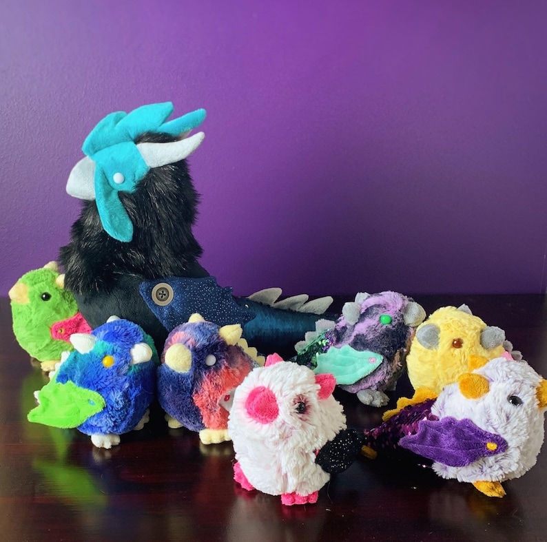 Pode incluir: Um grupo de brinquedos de pel&uacute;cia, incluindo um drag&atilde;o preto e azul, um p&aacute;ssaro verde e azul, um p&aacute;ssaro roxo e azul, um p&aacute;ssaro rosa e branco, um p&aacute;ssaro amarelo e um p&aacute;ssaro roxo e azul com um olho verde.