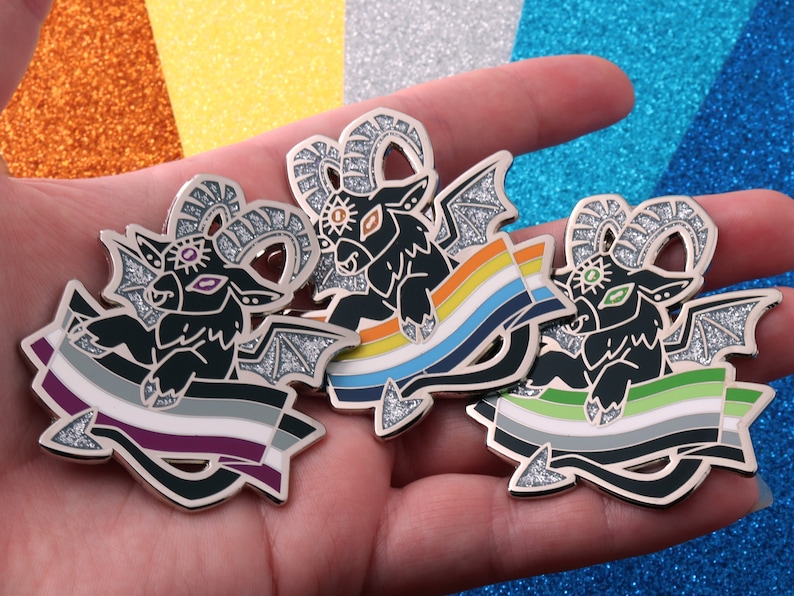 May include: Three enamel pins featuring a black, winged goat with a third eye and horns. Each goat is holding a different pride flag: asexual, bisexual, and pansexual.