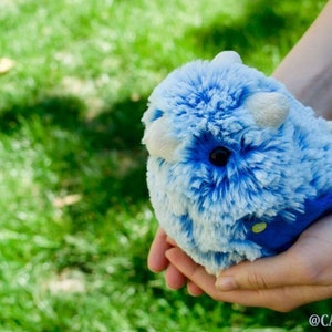 Pode incluir: Uma pessoa est&aacute; segurando um brinquedo de pel&uacute;cia azul e branco com um rosto branco e olhos pretos. O brinquedo est&aacute; vestindo uma roupa azul e verde.