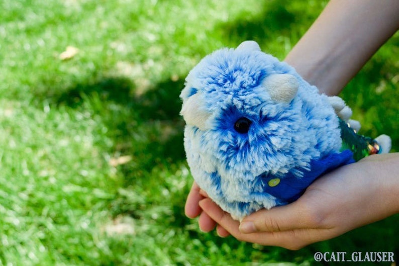 Pode incluir: Uma pessoa est&aacute; segurando um brinquedo de pel&uacute;cia azul e branco com um rosto branco e olhos pretos. O brinquedo est&aacute; vestindo uma roupa azul e verde.