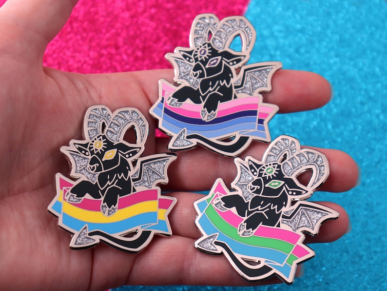 May include: Three black enamel pins with glitter accents, each featuring a three-eyed goat with wings and a banner. The banners are in the colors of the pansexual, bisexual, and transgender pride flags.