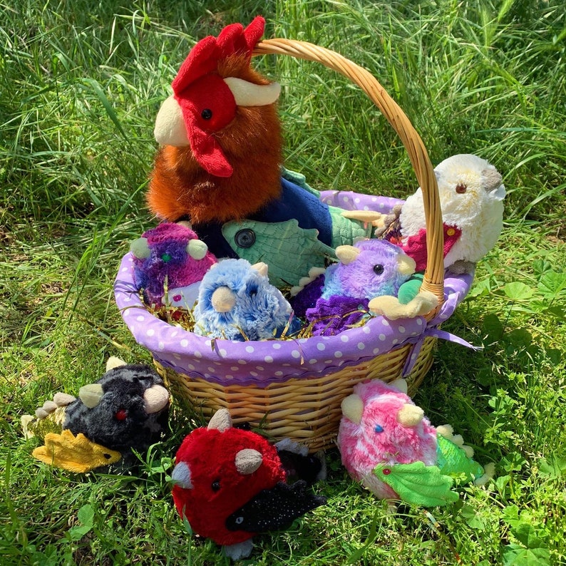 Pode incluir: Uma cesta de vime cheia de brinquedos de pel&uacute;cia coloridos, incluindo um galo, um drag&atilde;o e uma variedade de outras criaturas. A cesta &eacute; forrada com tecido roxo e tem bolinhas brancas.