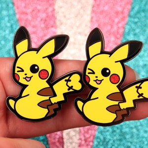 Puede incluir: Dos pines de esmalte de Pikachu de dibujos animados amarillos con contornos negros y mejillas rojas. Los pines están guiñando un ojo y tienen una cola marrón.