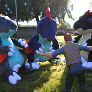 May include: Three plush toy dragons in blue, teal, and black with red accents. A person in a brown vest and blue jeans is standing in front of the dragons.