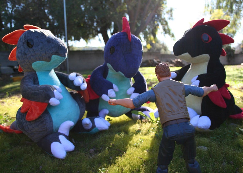 May include: Three plush toy dragons in blue, teal, and black with red accents. A person in a brown vest and blue jeans is standing in front of the dragons.