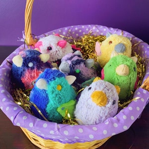 Pode incluir: Uma cesta de vime cheia de brinquedos de pel&uacute;cia coloridos. Os brinquedos s&atilde;o de v&aacute;rios formatos e tamanhos, incluindo um p&aacute;ssaro, uma flor e uma estrela. A cesta &eacute; forrada com tecido de bolinhas roxas e brancas e cheia de papel picado dourado e branco.